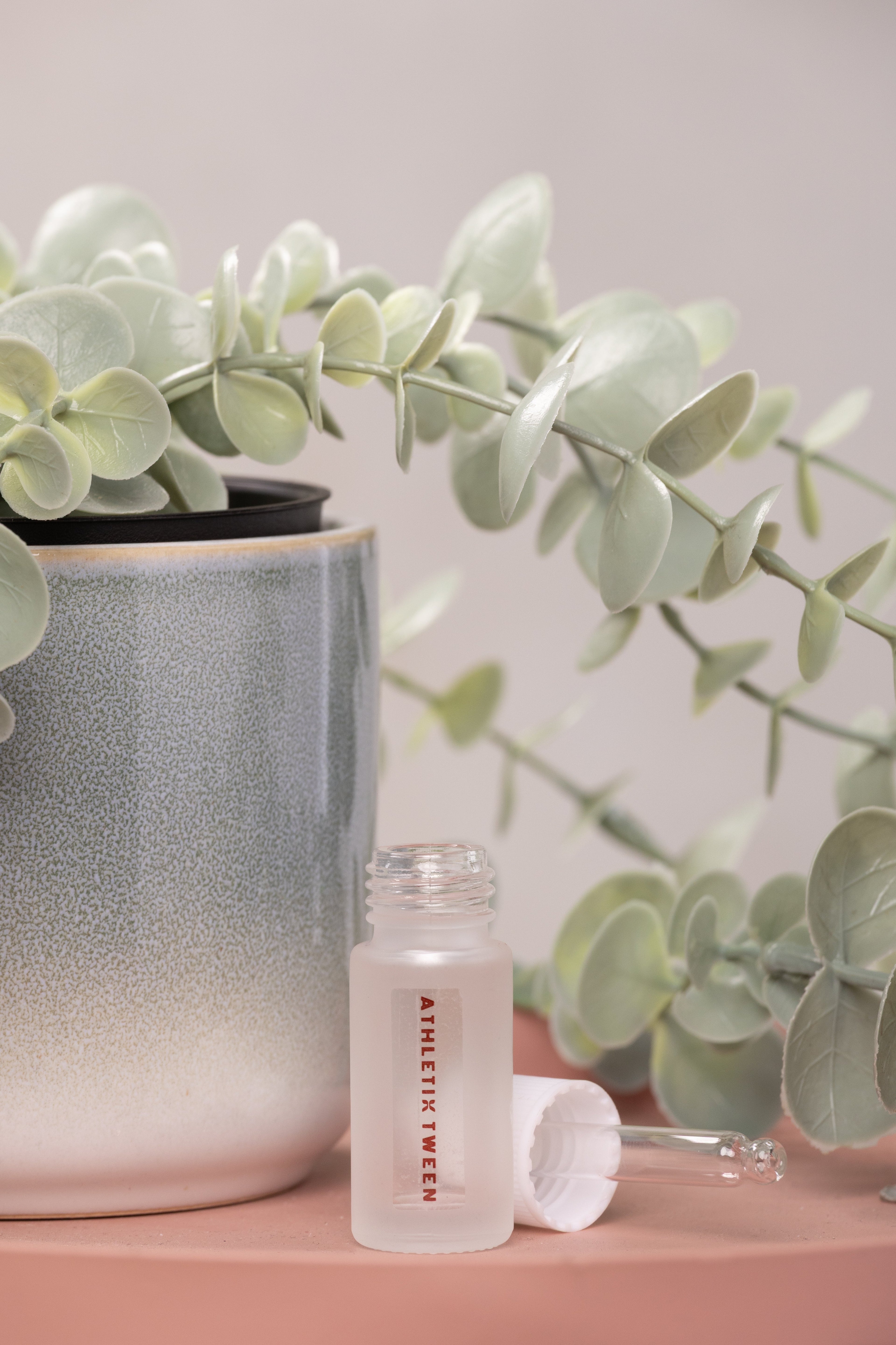 Small bottle labeled 'Athletic Tweed' with a plant in the background on a pink surface